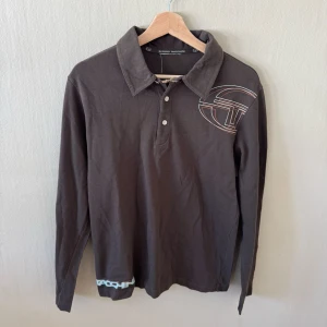 Sergio Tacchini Brun långärmad henley med tryck - Säljer en brun långärmad henley med tryck på bröstet och ett litet tryck vid nederkanten. Skjortan har klassisk krage, knappar framtill och är gjord i mjuk bomull. Perfekt för dig som gillar en avslappnad men ändå stilren look.0220
