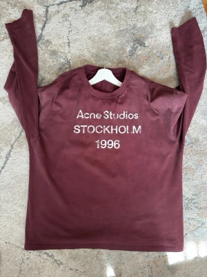 Vinröd långärmad topp Acne Studios - Vinröd långärmad topp från Acne Studios med tryck 'STOCKHOLM 1996' framtill. Rund halsringning och rak passform. Perfekt för dig som gillar stilrena och trendiga plagg med en touch av streetwear.