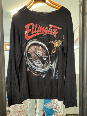 Svart långärmad topp Junk de Luxe - Cool svart långärmad topp från Junk de Luxe med stort tryck framtill i rött och vitt, samt motiv av klocka och skorpion. Rund halsringning och avslappnad passform. Perfekt för dig som gillar statement-plagg.