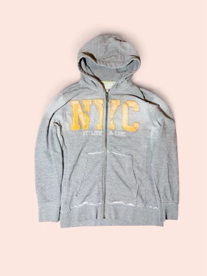 Grå hoodie med NYC-tryck - Säljer en grå zip-hoodie med stor huva och fickor framtill. På bröstet står det 'NYC' i stora orangea bokstäver. Perfekt för en chill och urban look.