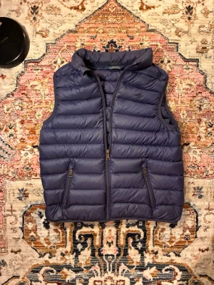 Mörkblå dunväst med hög krage - Snygg mörkblå dunväst med hög krage och två dragkedjeförsedda fickor framtill. Västen har quiltad design och stängs med dragkedja. Perfekt för lager-på-lager och håller dig varm under kyliga dagar. OBS det är en barnväst och vet inte vilken storlek det är men den är väldigt liten och skulle tro en xxxxs är kanske samma