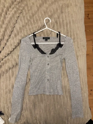 Grå ribbad långärmad topp Gina Tricot - Säljer en grå ribbad långärmad topp från Gina Tricot med knappar framtill och djup urringning. Toppen har en slim passform och är tillverkad i mjuk bomullsblandning. Perfekt att styla med ett spetslinne under för en snygg layering-effekt. skitsnygg ❤️