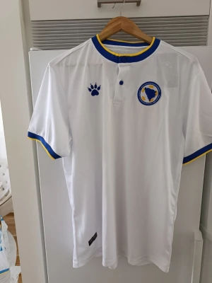Bosnien & Hercegovina vit pikétröja (Kelme) – Liljemönster - 🇧🇦 Snygg vit pikétröja för Bosnien & Hercegovina från Kelme! 🇧🇦 Perfekt inför matcher eller för dig som vill ha en riktigt stilren landslagströja. Tröjan är vit med blå och gula detaljer på krage och ärmar, knäppning i halsen, broderat förbundsmärke samt ett riktigt snyggt präglat mönster med bosniska liljor (Ljiljan) i tyget. Har fler storlekar.