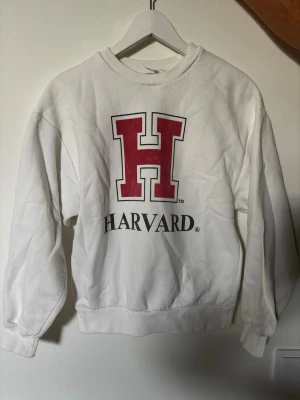 Vit sweatshirt med Harvard-tryck - Vit sweatshirt med stor röd H och texten Harvard på bröstet. Tröjan har rund halsringning, ribbade muddar vid ärmslut och nederkant. Perfekt för dig som gillar college-stil och vill ha en klassisk och bekväm tröja.