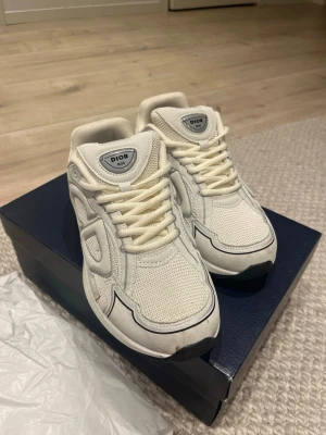 Vita Dior sneakers med meshdetaljer - Säljer ett par vita Dior sneakers med meshpaneler och snygga detaljer i läder och textil. Skorna har en chunky sula, vita snören och diskret logga på plösen. Perfekta för dig som vill ha en clean och exklusiv look.