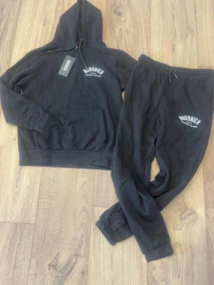 Hoodrich tracksuit (M) - Snyggt svart set från Modrich med hoodie och matchande byxor. Hoodien har ficka framtill och tryck på bröstet, byxorna har resår i midjan och tryck på ena benet. Perfekt för vardag eller mys! Storlek M. Både hoodie och byxor är i mjukt och bekvämt material.