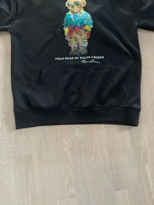 Svart hoodie Polo Bear Ralph Lauren - Svart hoodie från Ralph Lauren med ikoniska Polo Bear-trycket framtill. Tröjan har känguruficka och ribbade muddar. Perfekt för dig som gillar streetwear och vill ha en klassisk men lekfull look.