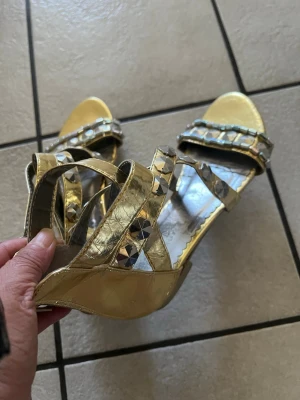 Guldiga sandaler med nitar storlek 37 - Säljer ett par glansiga guldiga sandaler med remmar och dekorativa silverfärgade nitar. Sandalerna har öppen tå och dragkedja bak för enkel påtagning. Perfekta för dig som vill sticka ut med metallic och coola detaljer.