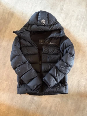 Sail racing vinterjacka - Snygg svart dunjacka med stor huva och quiltad design. Jackan har dragkedja framtill och logga på bröstet. Perfekt för kalla vinterdagar och håller dig varm med sin tjocka vaddering. Den har en snabbladdning på framsidan (bild 3) därav det låga priset(nypris lite mer än 3000kr). Den är perfekt på en kall vinterdag eller i skidbacken⛷️