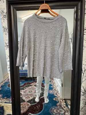 Grå stickad tröja med pärlor - Grå stickad tröja med rund hals och trekvartsärmar. Tröjan är dekorerad med små vita pärlor över hela framsidan för en trendig look. Materialet ser mjukt och bekvämt ut, perfekt för att lyfta din vardagsstil.