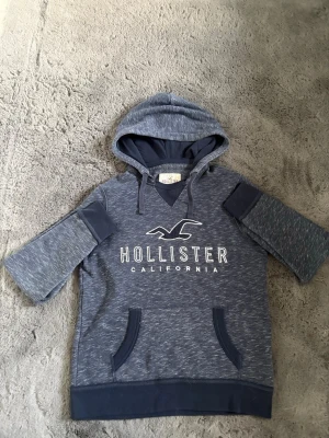 Blå Hollister hoodie med kort ärm - Snygg blåmelerad hoodie från Hollister med korta ärmar och huva. Stor logga och text 'Hollister California' framtill. Mörkare blå detaljer vid ärmslut, fickor och nederkant. Perfekt för en avslappnad och cool stil.