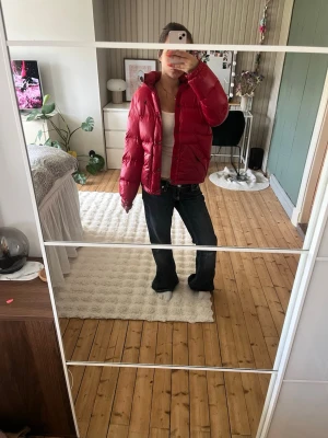 Ralph Lauren vinterjacka  - Säljer en riktigt snygg röd pufferjacka med huva och dragkedja samt knappar framtill. Jackan har två stora fickor och en liten logga på bröstet. Perfekt för kalla vinterdagar och ger en cool streetstil. Materialet är glansigt och fylligt för extra värme.