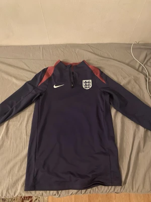 Mörkblå England Nike träningströja - Säljer en långärmad mörkblå träningströja från Nike med Englands landslagslogga på bröstet och röda detaljer på axlarna. Tröjan har halv dragkedja framtill och är perfekt för fotbollsträning eller uppvärmning.