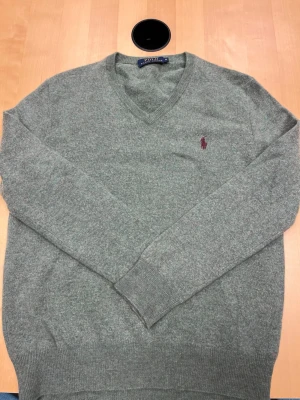 Grå stickad tröja Polo Ralph Lauren - Grå stickad tröja från Polo Ralph Lauren med V-ringning och lång ärm. Tröjan har en broderad vinröd logga på bröstet och ribbstickade muddar vid ärmslut och nederkant. Perfekt för en stilren och avslappnad look.
