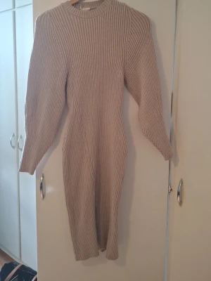 Beige ribbad stickad klänning - Säljer en beige ribbad stickad klänning med lång ärm och rund hals. Klänningen har en slits på sidan och är i en mjuk, stretchig kvalitet som sitter snyggt på kroppen. Perfekt för dig som gillar en enkel men trendig look.