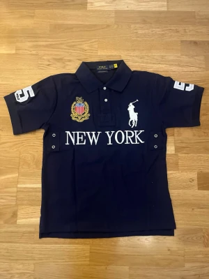 Mörkblå Ralph Lauren New York piké CHEIF KEEF edition - Snygg mörkblå pikétröja från Ralph Lauren ”CHEIF KEEF” med vit brodyr av logga och texten NEW YORK på bröstet. Har detaljer som broderad sköld, nummer 5 på ärmarna och klassisk krage med knappar. Perfekt för en sportig och stilren look. Storlek M, men jag skulle säga att den sitter mer som S. Skriv om du har några frågor.