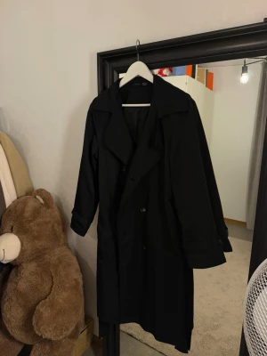 Svart trenchcoat - Stilren svart trenchcoat med bred krage och dubbelknäppning. Klassisk modell med långa ärmar och justerbara slejfar vid ärmsluten. Perfekt för dig som vill ha en tidlös och snygg jacka till hösten.