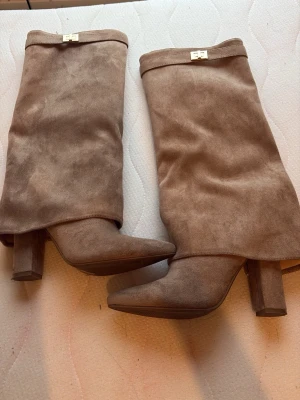 Beige höga stövlar i mocka - Säljer ett par snygga beige stövlar i mjuk mocka med spetsig tå och bred klack. Stövlarna är höga och har en stilren design som passar till många outfits. Perfekta för dig som vill ha ett par trendiga boots med modern känsla.