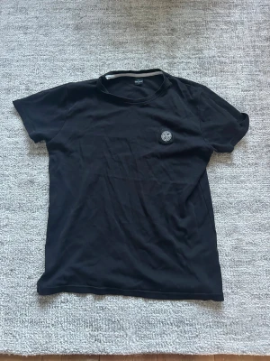 Svart t-shirt från Stone Island - Säljer en svart t-shirt från Stone Island med klassisk logga på bröstet. T-shirten har rund hals och korta ärmar, tillverkad i mjuk bomull för skön känsla. Perfekt till jeans eller shorts för en clean och stilren look.