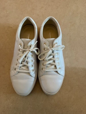 Vagabond vita låga sneakers i skinn - Stilrena vita sneakers med lågt skaft och klassisk design från vagabond i fint skick. Skorna har vita snören, guldfärgade öljetter och är tillverkade i slätt skinn. Perfekta för en clean och enkel look. Passar till det mesta och är lätta att matcha. Strl 40.