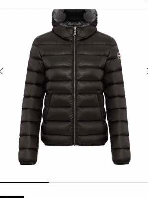 Svart dunjacka med huva - Snygg svart dunjacka med quiltad design och dragkedja framtill. Jackan har en hög krage och fast huva, samt en liten logotyp på ärmen. Perfekt för kalla vinterdagar och ger en stilren look. Nästan helt oanvänd. Säljer för jag inte passar i den. 