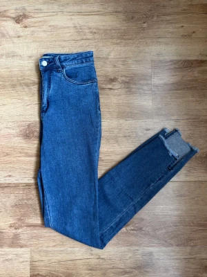 Blå skinny jeans med råa benslut - Snygga blå skinny jeans med hög midja och råa, avklippta benslut. Klassisk femficksmodell i stretchigt denimtyg som sitter tight hela vägen ner. Perfekta för en trendig och avslappnad look.