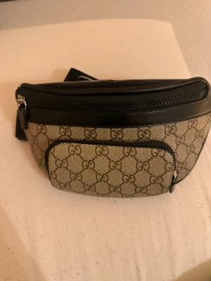 Magväska med GG-mönster - Snygg beige magväska med klassiskt GG-mönster och svarta läderdetaljer. Väskan har en justerbar rem, dragkedja upptill och en praktisk ytterficka framtill. Perfekt för dig som vill ha en trendig och ikonisk accessoar.