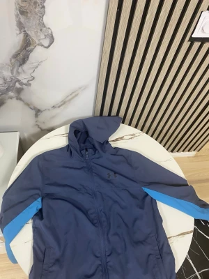 Under Armour vindjacka  - Snygg blå vindjacka från Under Armour med huva och dragkedja framtill. Jackan har ljusblå detaljer längs ärmarna och två fickor framtill. Perfekt för blåsiga dagar och sportiga outfits.