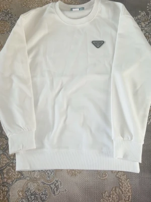 Vit sweatshirt från Prada - Snygg vit sweatshirt från Prada med klassisk rund halsringning och ribbade muddar vid ärmslut och nederkant. Tröjan har en ikonisk svart Prada-logga i metall på bröstet. Perfekt för en clean och stilren look.