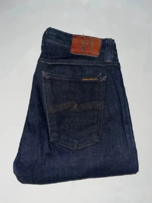 Nudie jeans  - Mörkblå Skinny Lin jeans från Nudie Jeans med smal passform och klassisk femficksdesign. Snygga kontrastsömmar och läderpatch baktill. Midja 35cm, längd 107cm. JAG SKICKAR EJ FLER BILDER NÄR JAG BÄR JEANSEN! för storleksguide hänvisar jag till måtten och för bättre inblick i passformen rekommenderar jag att googla på modellnamnet. kolla måtten noggrant :) (n1)