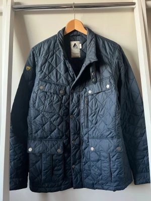 Mörkblå quiltad jacka från 8848 - Snygg mörkblå jacka med quiltat mönster och flera fickor framtill. Jackan har tryckknappar, dragkedja och hög krage med spänne. Materialet är lätt och vindtätt, perfekt för blåsiga dagar. Klassisk passform och stilren design.