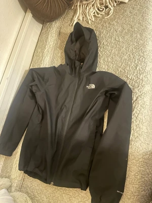 Svart regnjacka från The North Face - Svart regnjacka från The North Face med huva och hel dragkedja framtill. Jackan har logga på bröstet och är tillverkad i ett vattenavvisande material. Perfekt för regniga dagar och outdoor-äventyr.