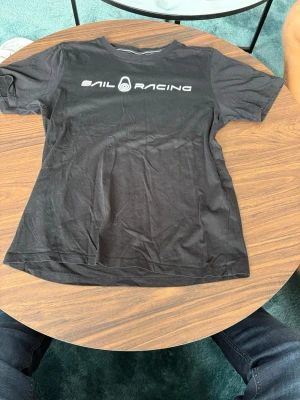 Svart Sail Racing t-shirt - Snygg svart t-shirt från Sail Racing med vit logga och text på bröstet. Klassisk rund halsringning och korta ärmar. Tillverkad i mjukt bomullsmaterial som känns skönt mot huden. Perfekt för dig som gillar stilrena och sportiga plagg.