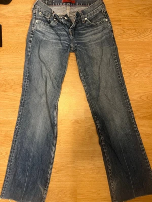 Blå bootcut jeans från Levi's - Säljer ett par klassiska blå bootcut jeans från Levi's med slitna detaljer. Jeansen har normal midja, dragkedja och knapp framtill samt den ikoniska läderpatchen bak i linningen. Perfekta för en avslappnad och trendig look.