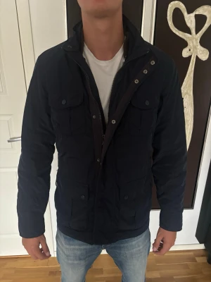 Field jacket - Snygg mörkblå jacka med fyra framfickor och hög krage. Jackan har dragkedja och knappar framtill samt långärmad design. Perfekt för dig som vill ha en stilren och praktisk jacka till vardags. Har storlek S men passar mig som är 190 cm lång
