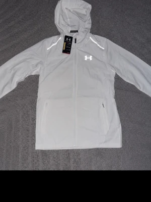 Vit vindjacka från Under Armour - Snygg vit vindjacka från Under Armour med huva och hel dragkedja framtill. Jackan har två sidofickor, reflekterande logga på bröstet och stor Under Armour-text på ryggen. Tillverkad i lätt och vindtätt material, perfekt för blåsiga dagar.
