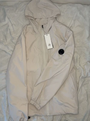 C.p company vindjacka - Snygg beige vindjacka med huva och dragkedja framtill. Jackan har en svart detalj på ärmen och elastiska muddar vid handlederna. Tillverkad i lätt och vindtätt material, perfekt för blåsiga dagar.
