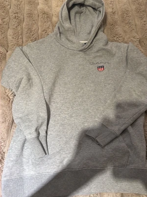 Grå hoodie från GANT med logga - Säljer en stilren grå hoodie från GANT med broderad logga och sköld på bröstet. Hoodien har huva, ribbade muddar och en klassisk passform. Perfekt för dig som gillar enkel och tidlös stil.