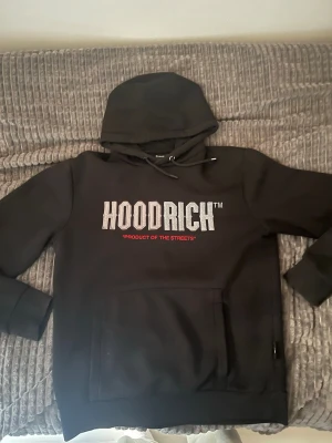 Svart HOODRICH hoodie med tryck - Svart hoodie från HOODRICH med stor vit logga och texten 'PRODUCT OF THE STREETS' i rött på bröstet. Klassisk känguruficka framtill och justerbar huva med snören. Perfekt för en streetwear-look.