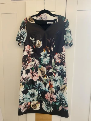 Blommig svart kortklänning - Snygg svart kortklänning med färgglatt blommönster i blått, rosa, beige och grönt. Klänningen har korta ärmar och en lätt v-ringad halsringning. Klänningen har också en dragkedja på baksidan. Perfekt för dig som vill sticka ut med en färgstark look.                                                               Är öppen för att förhandla priset🌸