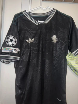 Juventus borta tröja med Thuram 19 - Snygg svart fotbollströja med diskret blommönster, silverfärgade detaljer och trycket Thuram 19 på ryggen. Tröjan har Champions League-märke på ärmen, randiga muddar och v-ringad krage. Tillverkad i lätt och ventilerande material.