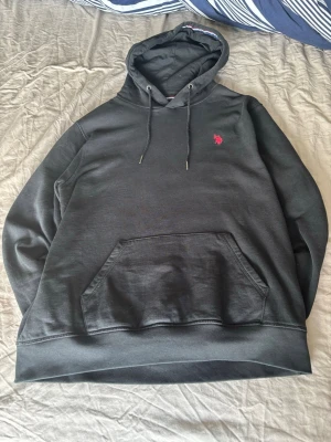 Svart hoodie med röd logga - Säljer en svart hoodie med huva och dragsnören. På bröstet finns en liten röd broderad logga. Hoodien har en stor magficka framtill och ribbade muddar vid ärmslut och nederkant. Perfekt för en avslappnad och stilren look.