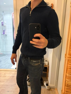 Mörkblå stickad tröja med dragkedja - Snygg mörkblå stickad tröja med halv dragkedja i halsen. Tröjan har ribbade muddar vid ärmslut och nederkant samt en hög krage som kan vikas ner. Perfekt för lager på lager och passar till jeans eller chinos.