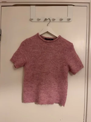 Rosa stickad t-shirt med kort ärm - Mysig rosa stickad t-shirt med korta ärmar och rund hals. Tröjan har en rak passform och är tillverkad i ett mjukt, melerat garn som ger en fluffig känsla. Perfekt för lager-på-lager och en avslappnad stil.