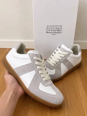 Maison Margiela GATs - Maison Margiela sneakers i vitt och ljusgrått med klassisk låg silhuett. Ovandel i slätt läder och mocka, vita snören och gummisula. Diskret Maison Margiela-logga på plösen. Perfekta för en clean och stilren look.