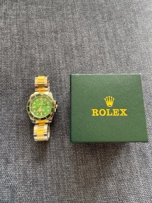 Rolle klocka - Snygg rolle klocka med grön urtavla, gulddetaljer och silverfärgad länk. Klockan har tydliga markeringar, datumvisning och klassisk rund form. Levereras i original grön box.