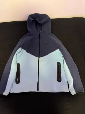 Nike hoodie i blått och ljusblått - Snygg hoodie från Nike i två nyanser av blått med dragkedja framtill och huva. Jackan har svarta detaljer vid fickorna och är tillverkad i ett mjukt material. Perfekt för en sportig och avslappnad stil.