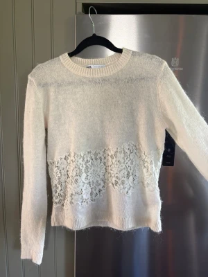Vit stickad tröja med spetsdetalj - Snygg vit stickad tröja med lång ärm och rund halsringning. Tröjan har en bred spetsdetalj med blommönster runt midjan. 💓 Perfekt att matcha med jeans eller kjol för en trendig look.