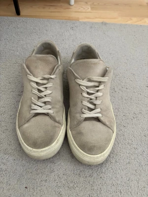 Beige mockasneakers med vit sula - Snygga sneakers i beige mocka med klassisk låg profil och vita snören. Skorna har en tjock vit gummisula och stilren design utan synliga loggor. Perfekta för dig som gillar minimalistisk och clean stil.