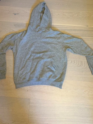 Grå hoodie med magficka - En enkel och stilren grå hoodie med huva och stor magficka framtill. Tillverkad i mjukt sweatshirtmaterial och har ribbade muddar vid ärmslut och nederkant. Perfekt för en avslappnad look.